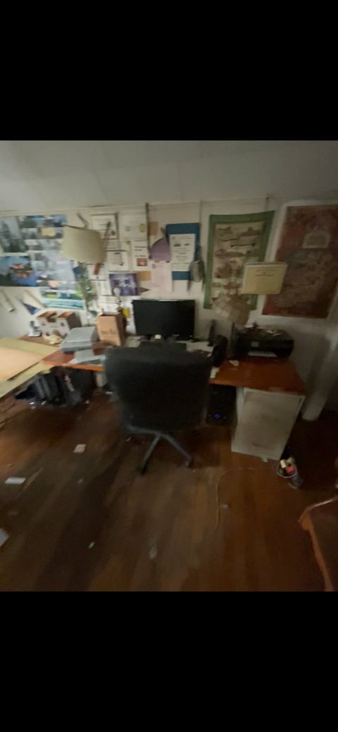 Cluttered home office with desk, computer monitor, office chair, and various papers and posters covering walls and floor