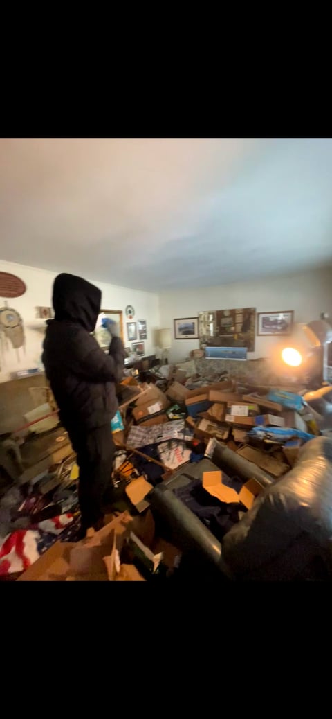 Person in black hoodie standing in cluttered living room filled with cardboard boxes and items, with warm lamp light and wall decorations