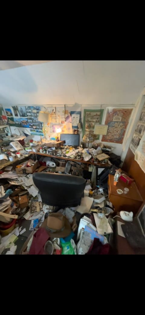 Cluttered bedroom with piles of items covering the floor, desk, and furniture, along with wall decorations and organized storage areas.