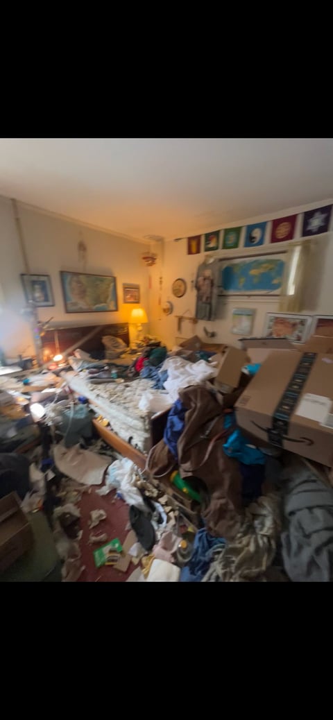 Messy bedroom with clothes scattered on bed and floor, desk with items, and wall decorations including colorful artwork and posters